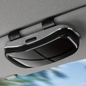 May include: A black and silver car visor sunglasses holder with a glossy finish and a rectangular shape. The device features a secure clip mechanism attached to a gray fabric vehicle sun visor, providing a compact storage solution for eyewear.