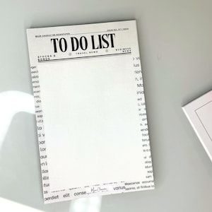 May include: A white notepad with the words "TO DO LIST" in bold, black letters. The notepad is designed to resemble a newspaper, with additional text such as "STOCKS & BONDS" and "TRAVEL NEWS". The notepad is on a light gray surface.