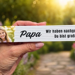 May include: A white folding ruler with the word "Papa" in black script and a photo of two children. The ruler also has the German text "Wir haben nachgemessen... Du bist großartig!"