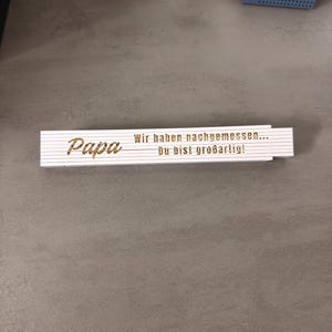 May include: A white and gold folding ruler with the word "Papa" and the German text "Wir haben nachgemessen... Du bist großartig!" printed on it. The ruler is lying on a grey surface.