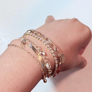 May include: A selection of gold-coloured bracelets. The bracelets include a chain link style, a tennis bracelet, a herringbone style, and a delicate chain with charms. The bracelets are layered on the wrist.