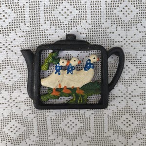 Könnte beinhalten: Ein schwarzer gusseiserner Topfuntersetzer in Teekannenform mit einem Design von drei Enten. Die Enten sind weiß mit orangefarbenen Füßen und Schnäbeln und tragen blau-weiß gepunktete Schleifen. Der Untersetzer befindet sich auf einem weißen Spitzengrund.