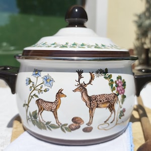 Puede incluir: Olla de esmalte blanco con tapa y asas marrones. La olla presenta un diseño decorativo de dos ciervos, flores azules y uvas moradas. Adecuada para cocinar y servir.