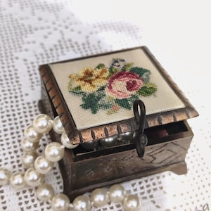 Könnte beinhalten: Kleine, antike Holzschmuckschatulle mit floralem Nadelspitzendeckel, geöffnet, um eine Perlenkette zu enthüllen. Die Schatulle ist braun mit dekorativem Metallverschluss und Zierleisten. Die Perlen sind weiß und über die Schatulle drapiert.