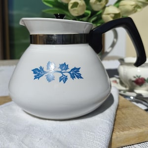 Peut inclure: Théière en verre blanc avec une anse et un couvercle noirs. La théière présente un motif de feuilles bleues et une bande argentée. Un tissu blanc et une planche à découper en bois sont au premier plan.