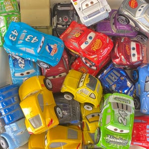 Op de afbeelding: Een verzameling speelgoed raceauto's in verschillende kleuren, waaronder rood, blauw, geel en groen. De auto's hebben verschillende ontwerpen en nummers, sommige met de logo's "Rust-eze" en "Dinoco". De auto's zitten in een witte container.