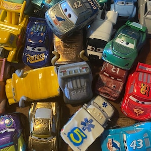 May include: A collection of various die-cast toy cars featuring characters from the Cars movie franchise. The assortment includes vehicles in red, blue, yellow, green, purple, and gold, displaying racing numbers, logos, and expressive eyes.