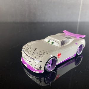 Op de afbeelding: Een speelgoedauto met een lichtgrijze carrosserie met zwarte spikkels, een paarse voorbumper, paarse wielen en een paarse achterspoiler. De auto heeft groene ogen op de voorruit, een glimlachende mond op de grille en het nummer 012 in rood.