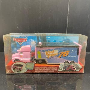 Op de afbeelding: Een roze Disney Pixar Cars speelgoed vrachtwagen en trailer set. De trailer is blauw met de tekst "Vinyl Toupee Racing 76" en sterren graphics. De set zit in een doorzichtige plastic display case.