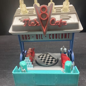 Può includere: Una replica giocattolo del Flo's V8 Cafe del film Cars. Il modello presenta una base turchese, un tetto grigio con dettagli in plastica trasparente e un'insegna rossa e grigia con la scritta "Flo's Cafe". L'insegna ha anche un logo "8".