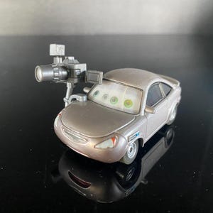 Op de afbeelding: Een zilverkleurige speelgoedauto met een metallic afwerking en een cameraset gemonteerd op de motorkap. De auto heeft een voorruit met geprinte ogen, een grille en koplampen. De camera-uitrusting bestaat uit een lens, zoeker en een klein scherm.