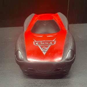 Op de afbeelding: Een rood en zwarte speelgoedauto met het woord "Cars" en het nummer "2" op de voorkant. De speelgoedauto heeft zwarte koplampen en een zwarte voorbumper. De auto staat op een donkere ondergrond.