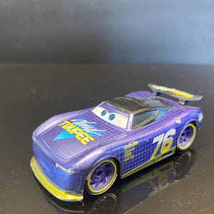 Op de afbeelding: Een paarse speelgoed raceauto met het nummer 76 aan de zijkant. De auto heeft gele accenten op de wielen en de spoiler. De tekst "Wheel Toupee" is zichtbaar op de motorkap. De auto heeft blauwe ogen.
