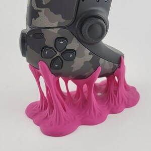 May include: A camouflage patterned video game controller resting on a bright pink, organic-shaped stand. The stand features a textured, dripping, or web-like design that supports the base of the controller, which includes a directional pad and joystick.