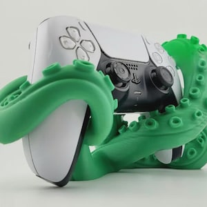 Puede incluir: Un mando de videojuegos blanco y negro sostenido por un soporte con forma de tentáculo de pulpo de color verde brillante. El mando cuenta con un panel táctil central, dos joysticks analógicos y botones direccionales. La base tiene ventosas.