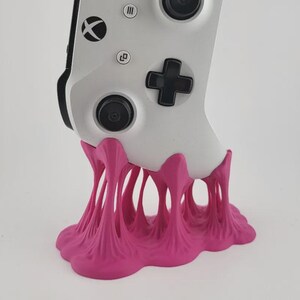 May include: A white Xbox video game controller resting in a bright pink, 3D-printed stand. The stand features a unique, organic, melted-look design with multiple vertical supports that connect to a wide, irregular base, holding the controller upright.