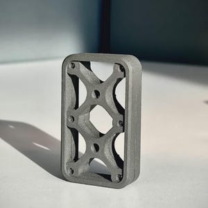 May include: A rectangular, dark gray, matte-finished object with a thick outer frame and an internal structural lattice featuring four circular openings and a central diamond-shaped void, constructed with a textured, granular surface appearance.