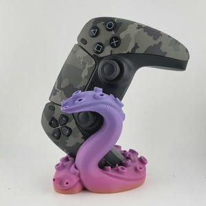以下が含まれることがあります： グレーの迷彩柄のビデオゲームコントローラーが、紫色のタコの触手スタンドに置かれています。コントローラーには標準的な黒いボタン、ジョイスティック、方向パッドが付いています。触手のベース部分には吸盤がはっきりと見えます。