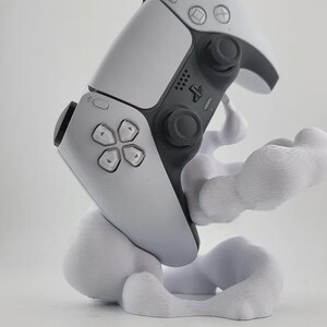 May include: A white and black video game controller resting on a white, cloud-shaped stand. The controller features a directional pad, two analogue sticks, and the PlayStation logo. The stand has a textured, layered appearance with rounded, bulbous forms.