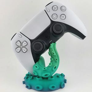 Könnte beinhalten: Ein weiß-schwarzer kabelloser Videospiel-Controller auf einem türkisfarbenen Oktopus-Tentakel-Ständer. Der Ständer verfügt über eine kreisförmige Basis mit mehreren Saugnäpfen und einen geschwungenen Tentakelarm, der den Controller aufrecht hält.