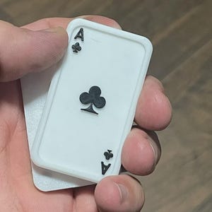 May include: A white rectangular playing card with a raised black club symbol in the centre and the letter A with a small club symbol in the top left and bottom right corners. The card features a thick white border and a textured surface finish.