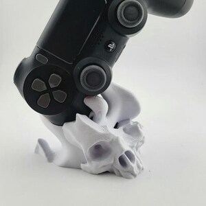 以下が含まれることがあります： スタイライズされた頭蓋骨の形をした白い3Dプリントスタンドに置かれた黒いビデオゲームコントローラー。コントローラーには中央にPlayStationロゴボタン、2つのアナログスティック、方向パッドがあり、ベース部分は質感のある表面になっています。