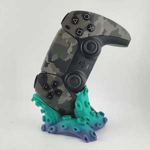 以下が含まれることがあります： カモフラージュ柄のビデオゲームコントローラーが、ティールとパープルのタコの触手型スタンドに置かれています。コントローラーには黒いボタン、ジョイスティック、方向パッドがあり、グレー、ブラック、オリーブグリーンの迷彩柄のシェルが特徴です。