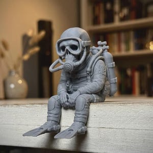May include: A grey monochromatic figurine of a skeleton wearing a full scuba diving suit, including a mask, regulator, and oxygen tank. The figure is depicted in a seated position with legs dangling, wearing flippers on its feet.