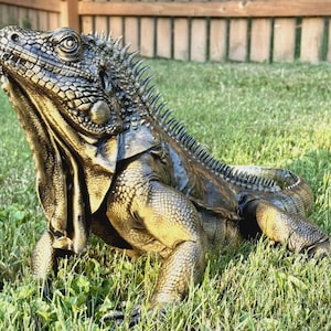 Puede incluir: Una escultura de iguana de color bronce con escamas muy detalladas, una papada prominente bajo la barbilla y una hilera de espinas afiladas a lo largo del lomo. La figura del lagarto se muestra en una pose realista con la cabeza levantada.