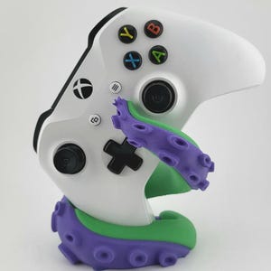 May include: A white Xbox wireless controller resting on a purple and green octopus tentacle stand. The controller features a black D-pad, black analogue sticks, and four coloured action buttons labelled A, B, X, and Y. The stand has suction cup details.