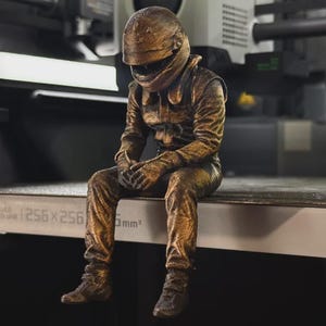 May include: A bronze-coloured figurine of a race car driver wearing a full-face helmet and a racing suit, sitting in a hunched posture with hands clasped. The figure has a textured, metallic finish and is positioned on a flat surface.