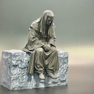 May include: A gray monochromatic figurine of a seated figure wearing a hooded robe with intricate textured patterns on the sleeves and hem. The figure sits with hands clasped on a rectangular block base that features a rough, stone-like surface texture.