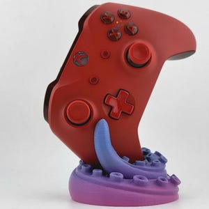 Könnte beinhalten: Ein roter Videospiel-Controller, der in einem violetten Oktopus-Tentakel-Ständer ruht. Der Controller verfügt über ein Steuerkreuz, zwei Analogsticks und vier Aktionstasten mit der Beschriftung A, B, X und Y. Die Tentakelbasis hat Saugnäpfe.