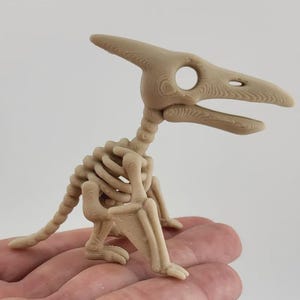 Op de afbeelding: Een kleine, lichtbruine, 3D-geprinte Pterodactylus-skeletfiguur. Het model heeft een gedetailleerde schedel met een grote oogkas, een gesegmenteerde nek, een ribbenkast, een lange staart en beweegbare vleugels met zichtbare lijnen.