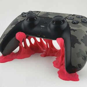 May include: A black and gray camouflage patterned video game controller resting on a bright pink, melted-look stand. The controller features standard black analog sticks, a central PlayStation logo, and action buttons with geometric symbols.