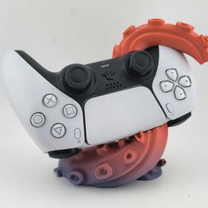 Könnte beinhalten: Ein weiß-schwarzer Videospiel-Controller auf einem dekorativen Ständer in Form eines gewundenen Krakenarms. Die Basis des Tentakels weist einen Farbverlauf von einem dunklen Violett an der Unterseite bis zu einem leuchtenden Orange auf.