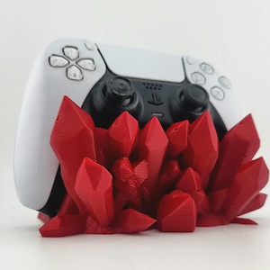 May include: A white and black video game controller resting in a red, 3D-printed stand shaped like a cluster of jagged crystals. The controller features a central black panel with the PlayStation logo and standard directional buttons and joysticks.