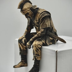 Puede incluir: Una figura dorada y negra de un soldado sentado con casco y armadura de estilo romano. La figura se muestra en una postura de descanso encorvada con una espada sujeta al cinturón, presentando un acabado texturizado y desgastado.
