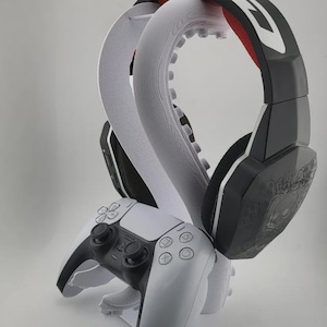Pode incluir: Um suporte branco impresso em 3D em forma de polvo que segura auscultadores de jogo pretos e brancos e um comando de jogo sem fios branco. O suporte apresenta tentáculos texturizados que se curvam para cima para apoiar o headset, enquanto o comando repousa por baixo.