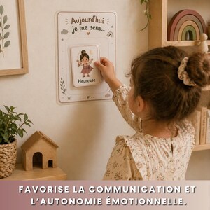 May include: A child selects a card from a wall-mounted board. The board reads "Aujourd'hui je me sens..." and features a card with a cartoon girl labeled "Heureuse." The image promotes emotional communication and autonomy. A wooden house and a rainbow are visible.