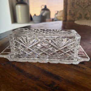 May include: A clear glass butter dish featuring a rectangular base and a matching lid. The exterior of the lid is decorated with a pressed diamond pattern and starburst motifs, while the base has a smooth, flat rim and a recessed center area.