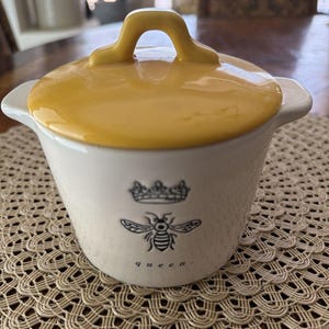 May include: A white ceramic lidded sugar bowl with a bright yellow lid featuring a handle. The front of the bowl displays a black line drawing of a crown above a bee, with the lowercase text "queen." printed in a simple font below the bee.