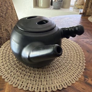 May include: A black ceramic teapot with a round body, a lid with two holes, and a handle. The teapot has a spout and sits on a woven placemat. The teapot is on a wooden table.