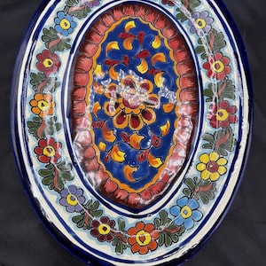 Puede incluir: Plato ovalado de cerámica con un vibrante diseño floral. El plato presenta un borde azul, un borde interior blanco y un diseño central en tonos de azul, rojo, amarillo y naranja. El patrón floral incluye varias flores coloridas.