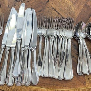 May include: A large collection of stainless steel flatware including multiple forks, knives, and spoons. The set features a simple, polished design with rounded handles, arranged in organized groups on a wooden surface.