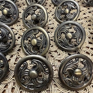 May include: A collection of round metal cabinet knobs featuring an antique brass finish. Each knob has a circular outer frame with an openwork design of swirling, curved scrollwork patterns and a central raised circular button detail.
