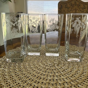 May include: Four clear glass tumblers with a square base, each featuring a white etched design of a flute-playing figure, spiral patterns, and cross symbols. The glasses have a tall, straight-sided shape with a smooth, transparent finish.