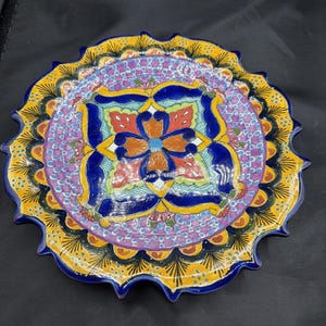 May include: A decorative ceramic plate with a vibrant, hand-painted design. The plate features a central floral motif in orange, blue, and red, surrounded by a purple and yellow border. The scalloped edge is outlined in blue. A colorful piece for home decor.