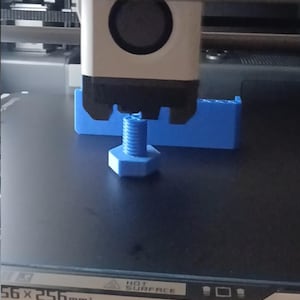 Puede incluir: Un perno azul claro impreso en 3D está siendo creado por una impresora 3D. El perno es hexagonal con un vástago roscado. También se ve una estructura de soporte azul. El cabezal de la impresora es blanco y negro.