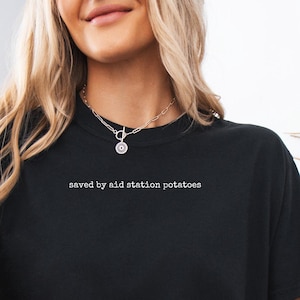 May include: A black short-sleeved t-shirt featuring the white text "saved by aid station potatoes" printed in a small font across the upper chest area. The shirt is paired with light blue denim shorts and a beige textured cardigan.